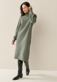 Abito maglione lavorato a maglia in verde oliva, con maniche lunghe a coste, collo alto e silhouette dritta, abbinato a stivali neri al ginocchio.