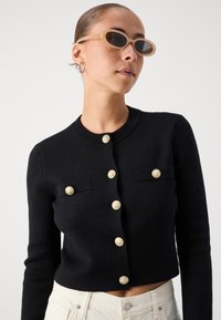 Black cropped cardigan with long sleeves, decorative gold buttons, and front pockets; textured knit fabric. Model wearing tan sunglasses.