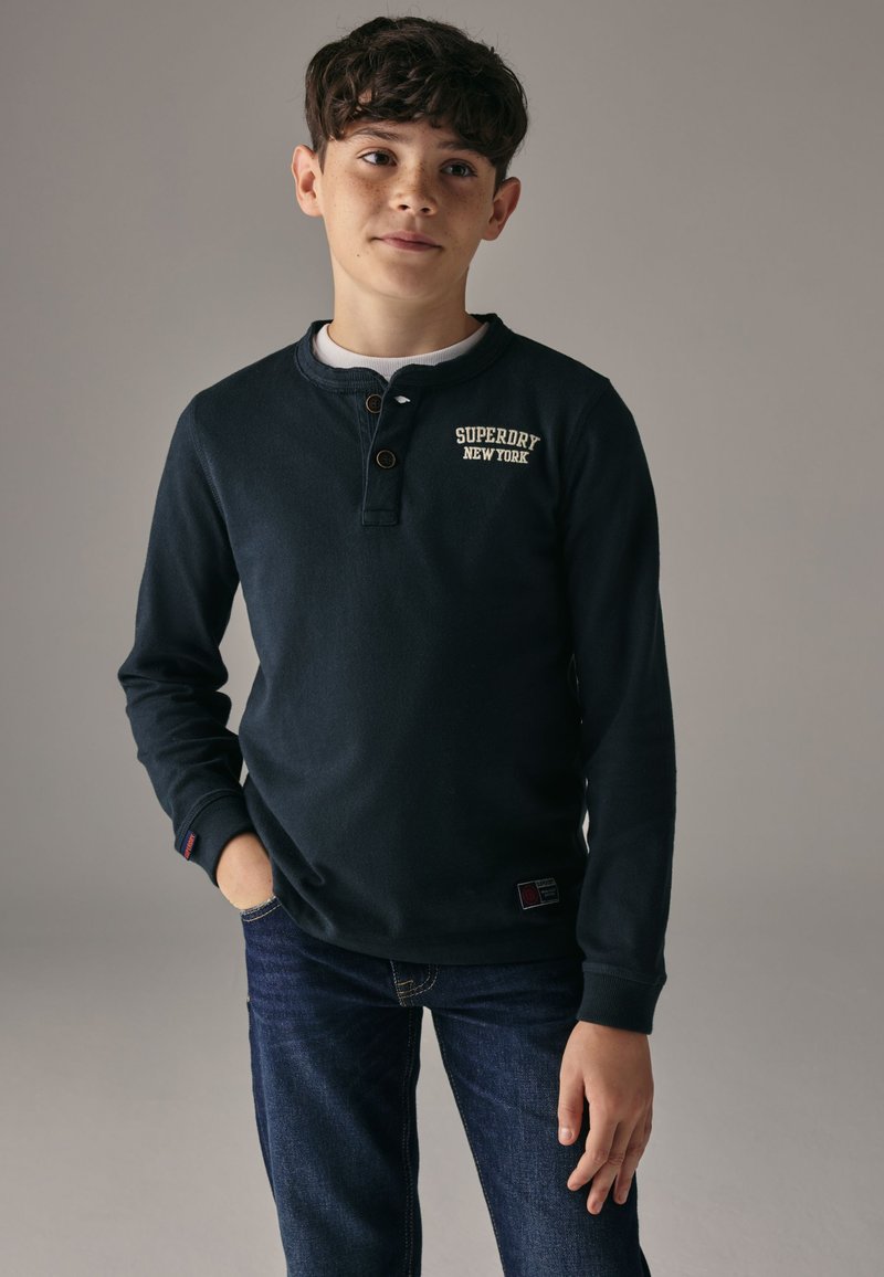 Mörk marinblå långärmad tröja i mjukt tyg med rund halsringning, knappdetalj och "Superdry New York"-logotyp i vitt. Kombinerad med jeans.
