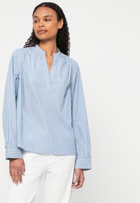 Blå og hvid stribet bluse med lange ærmer og stående krave, der har en afslappet pasform og manchetærmer. Glat stofstruktur.