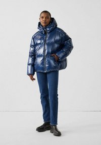 Giacca puffer blu navy con finitura lucida, dotata di cappuccio, chiusura con zip e polsini elasticizzati, abbinata a jeans in denim e scarponi neri.