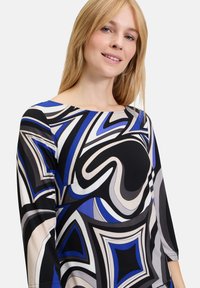 Bunt gemustertes Oberteil mit kräftigen schwarzen, blauen und beigen Wirbeln. Lange Ärmel und ein weiter Halsausschnitt, aus strukturiertem Stoff gefertigt.
