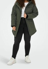 Olivgrön vadderad parka med huva, zip-fickor och svarta muddar, tillsammans med svarta skinny jeans och vita sneakers.