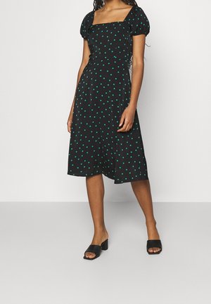 Femme portant une robe noire mi-longue à pois verts, avec un décolleté carré, des manches bouffantes et des chaussures noires à talons blocs ouverts à l'avant.