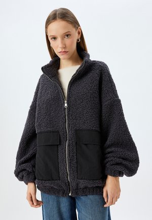 Mørkegrå fleecejakke med høj krave, frontlynlås og sorte stoflommer. Har voluminøse ærmer og et struktureret, hyggeligt udseende.