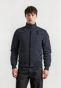 Young man wearing a zipped navy bomber jacket with ribbed cuffs and collar, standing against a plain light background.
