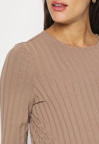 Blusa de manga longa castanho claro com textura canelada e decote redondo. Apresenta riscas verticais subtis e um silhueta ajustada.