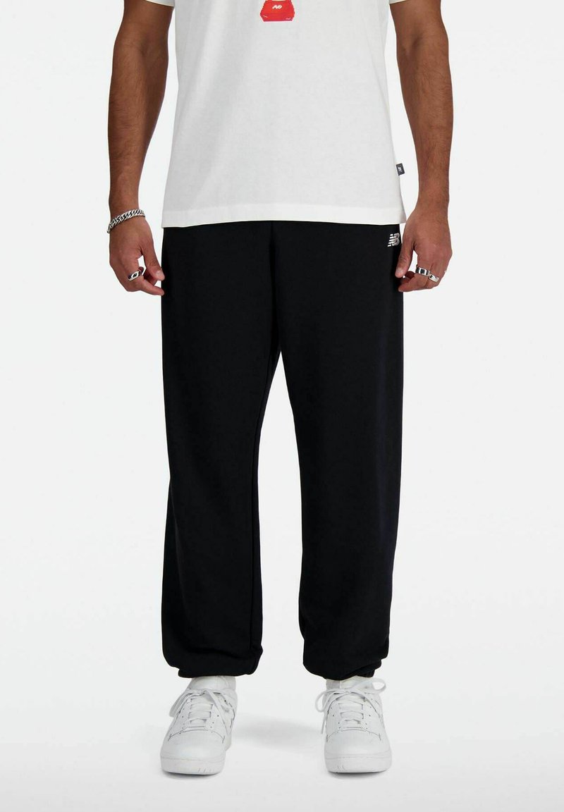 Schwarze Jogginghose aus weichem Stoff, lockere Passform, mit einem weißen Logo auf dem linken Bein, kombiniert mit weißen Sneakers und einem schlichten weißen Shirt.