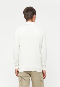 Hombre joven visto de espaldas vistiendo un suéter blanco de manga larga y pantalones cargo beige frente a un fondo blanco liso.