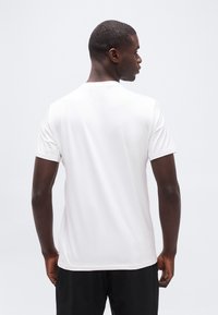 Άνδρας που φοράει απλό λευκό κοντομάνικο μπλουζάκι και μαύρο παντελόνι, στέκεται με την πλάτη στην κάμερα μπροστά σε λευκό φόντο.