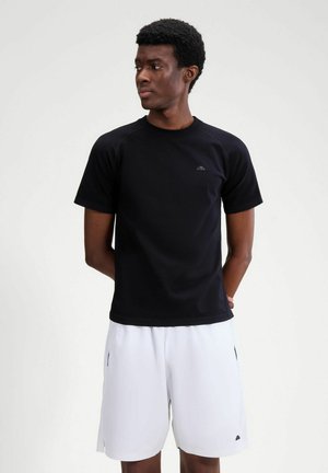 Junger Mann steht mit den Händen hinter dem Rücken, trägt ein schwarzes kurzärmeliges Hemd und weiße knielange Shorts vor einem schlichten Hintergrund.