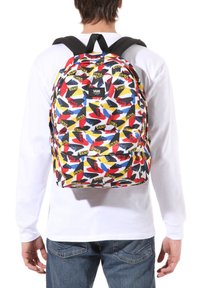 Mochila Vans multicolor con un patrón de logotipos repetidos en rojo, amarillo y azul sobre un fondo blanco, con correas ajustables negras.