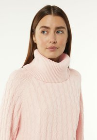 Helle rosa, gestrickte Pullover mit einem gerippten, oversized Rollkragen und einem strukturierten Muster, das erhabene, sich miteinander verflechtende Formen zeigt.