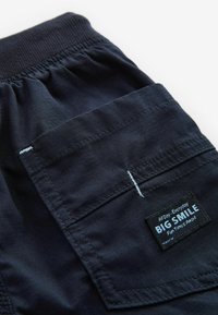 Marineblå shorts med elastisk talje, prydet med en stor lappelomme og et sort mærke med teksten "BIG SMILE" i hvid.