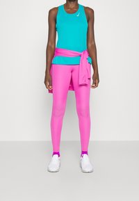 Débardeur de sport turquoise, leggings roses et baskets blanches. Les leggings ont une texture lisse, le débardeur arbore un logo, et un pull noué en guise d'accent.
