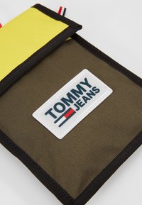 Borsa rettangolare in canvas marrone con un cappuccio giallo. Caratterizzata da una toppa con logo bianco etichettata "TOMMY JEANS" e rifiniture nere lungo i bordi.