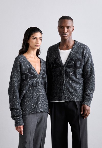 Dark gray, textured knit cardigans with a speckled pattern and bold black lettering, featuring button front and ribbed cuffs, worn over gray trousers.