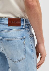 Jeans in denim celeste con una toppa in pelle marrone che mostra la scritta "BOSS." Presenta due tasche posteriori e una leggera scoloritura sul tessuto.