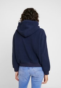 Felpa con cappuccio blu navy in pile, dal design ampio, con polsini e orlo elasticizzati, e una texture morbida, indossata con jeans azzurri.