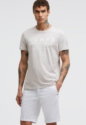 Mann mit lockigem Haar und Tattoos, der ein hellbeiges T-Shirt mit Aufdruck und weiße Shorts trägt, mit einer Hand in der Tasche.