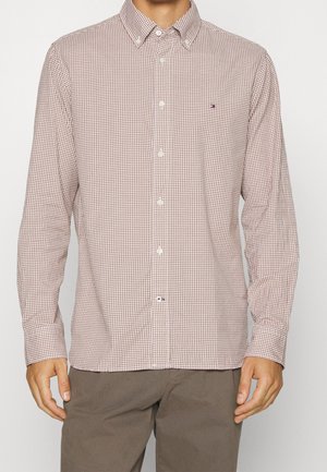 Uomo che indossa una camicia a maniche lunghe beige e marrone con piccoli quadretti e pantaloni marroni, in piedi davanti a uno sfondo uniforme.