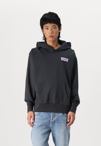 Sudadera negra con capucha y un bolsillo frontal, con un logo morado de Levi's en el pecho y puños acanalados. Se lleva con jeans de mezclilla azul claros.
