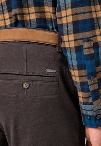 Pantaloni marroni in tessuto testurizzato, con una cintura in camoscio beige e una tasca posteriore con bottone, con un'etichetta di marca visibile. Camicia a scacchi blu e gialla.