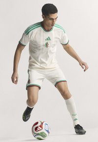 White soccer kit with green and red accents features Algeria crest, Adidas logo, and black soccer cleats near a multicolored ball.