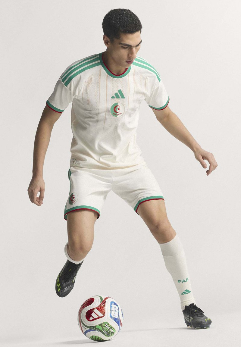 White soccer kit with green and red accents features Algeria crest, Adidas logo, and black soccer cleats near a multicolored ball.