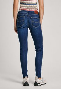 Kvinna i mörkblå tajta jeans, randig ärmlös topp och vita sneakers, som står vänd bort från kameran mot en enfärgad bakgrund.