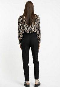 Kobieta z długimi, prostymi brązowymi włosami, ubrana w czarne spodnie oraz czarną, wzorzystą bluzkę z długim rękawem, stojąca tyłem do kamery.