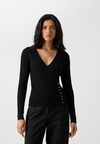 Haut enveloppant à manches longues en tricot noir côtelé avec un décolleté en V et des boutons en argent décoratifs sur le côté gauche. S'ajuste parfaitement au corps.