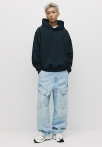 Tengerészkék kapucnis pulóver kenguruzsebbel, világoskék cargos farmernadrággal, amelyen több zseb található. Ezüst sportcipővel viselve.