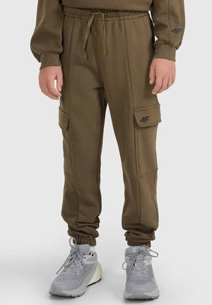 Osoba ubrana w oliwkowe spodnie joggery typu cargo i szare buty sportowe, stojąca na jednolitym tle.