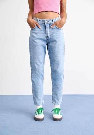 Personne portant un jean taille haute bleu clair, un crop top rose, des chaussettes blanches et des baskets blanches avec des rayures et des lacets verts, debout sur un sol bleu.