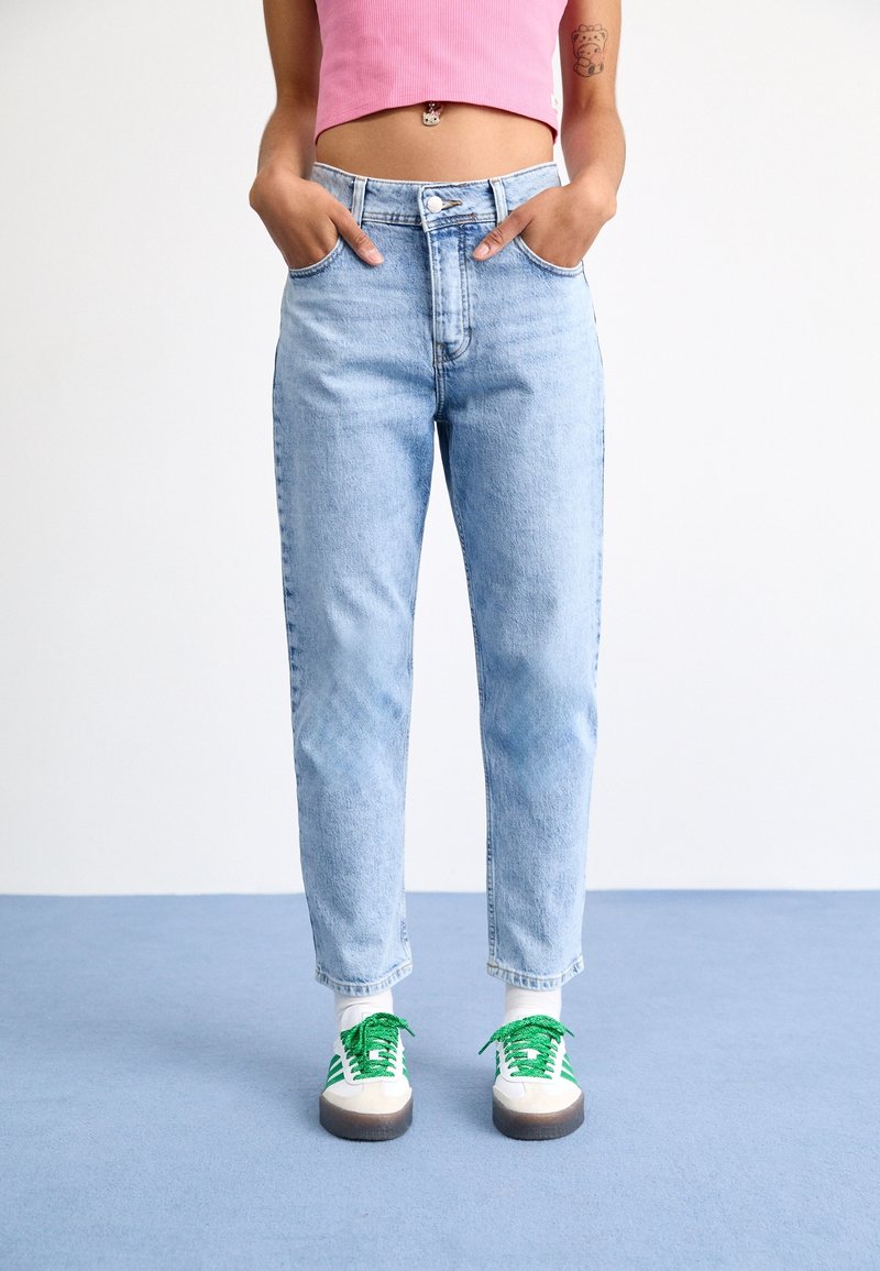 Personne portant un jean taille haute bleu clair, un crop top rose, des chaussettes blanches et des baskets blanches avec des rayures et des lacets verts, debout sur un sol bleu.