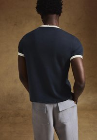 Tengerészkék rövid ujjú póló fehér szegéllyel, világosszürke nadrággal, amelyen két foltzseb található. Texturált háttér.