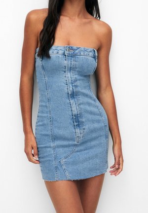 Mini sukienka z jasnoniebieskiego denimu bez ramiączek, z przodu zdobiona guzikami i przeszyciami, noszona przez stojącą osobę o długich, ciemnych włosach.