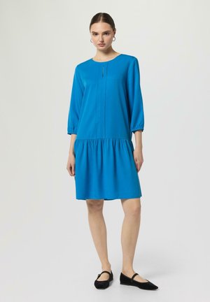 Frau steht in einem leuchtend blauen knielangen Kleid mit Dreiviertelärmeln und schwarzen flachen Schuhen vor einem schlichten hellen Hintergrund.