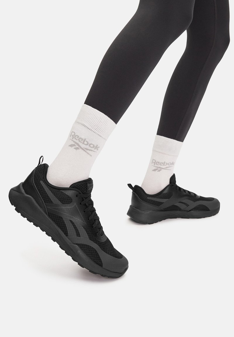 Juodi sportiniai batai su tinklinio tipo viršumi, guminiu padu ir tekstūruotais akcentais. Dėvimi su baltais kojinėmis, kuriose yra pilkas logotipas.