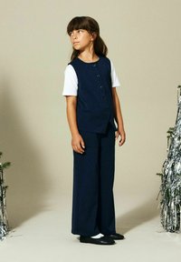 Ung jente står innendørs, iført marineblå knappet vest, vide bukser, hvit t-skjorte og svarte ballerinasko, og ser mot siden.