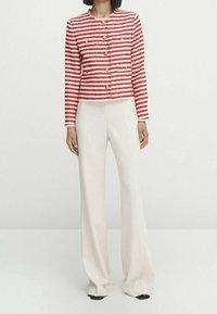 Veste en tweed rayée rouge et blanche avec des boutons dorés, associée à un pantalon blanc à jambes larges. Texture lisse et coupe courte.