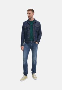 Chaqueta de denim de color azul oscuro, con dos bolsillos en el pecho; camiseta verde debajo, jeans de color azul claro y zapatillas crema con cordones blancos.