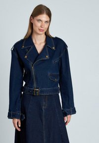 Chaqueta de mezclilla con cintura ceñida, cierre delantero con cremallera y múltiples bolsillos. Color azul oscuro, costuras prominentes y diseño de solapa ancha.