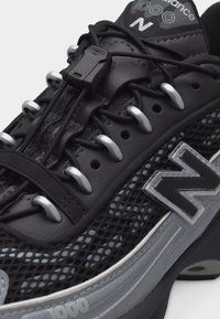 Μαύρο και γκρι αθλητικό παπούτσι New Balance με ύφασμα από δίχτυ, σύστημα γρήγορου κλειδώματος κορδονιών και λογότυπο στο πλάι και τη γλώσσα.