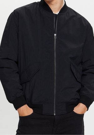 Homme portant une veste bomber noire zippée avec poches à rabat et poignets, col et taille côtelés, mains dans les poches.