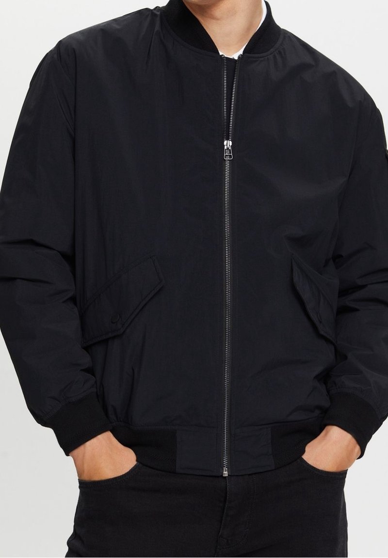 Homme portant une veste bomber noire zippée avec poches à rabat et poignets, col et taille côtelés, mains dans les poches.