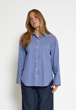 Žena s dlhými blond vlasmi, ktorá má na sebe modrú a bielu košeľu s vertikálnymi pruhmi a tmavé široké nohavice, stojí pred jednoduchým pozadím.