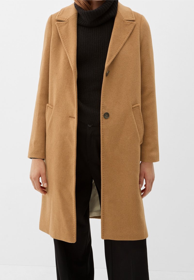 Persona che indossa un cappotto di lana color cammello sopra un maglione a dolcevita nero e pantaloni neri, in piedi di fronte a uno sfondo bianco semplice.