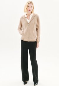 Sudadera beige con cremallera, hecha de tela suave, con capucha y bolsillos tipo canguro, combinada con pantalones de pierna ancha negros.